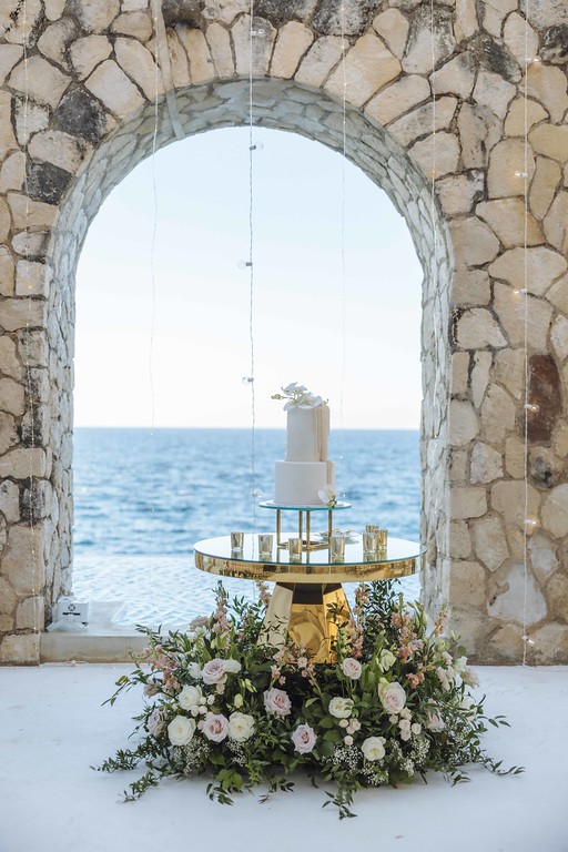 Wedding cake on a gold table framed by a stone arch overlooking the sea